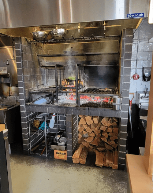A commercial kitchen grill with smoke, charcoal, and stacked firewood, showcasing a cooking area.