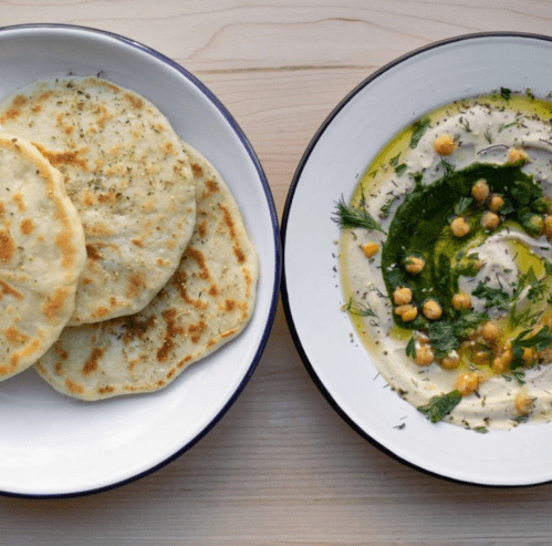 Three flatbreads on a plate next to a bowl of hummus topped with herbs, olive oil, and chickpeas.