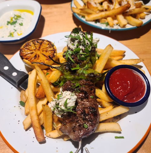 A plate of grilled meat skewers with fries, a charred lemon, and ketchup on the side, garnished with herbs.