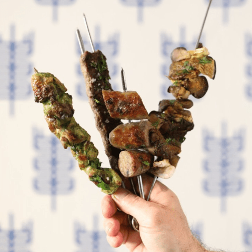 A hand holding four skewers of grilled meat and vegetables against a blurred background.