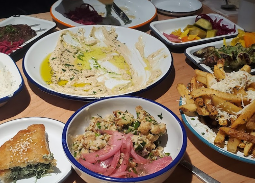 A table spread with various dishes, including hummus, fries, baklava, and colorful salads.