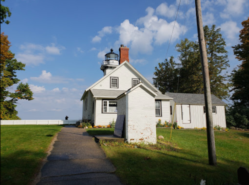 A white lighthouse beside a house, surrounded by trees and a clear blue sky with scattered clouds.