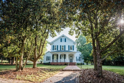 A charming white house with green shutters, framed by trees and a pathway leading to the front porch.