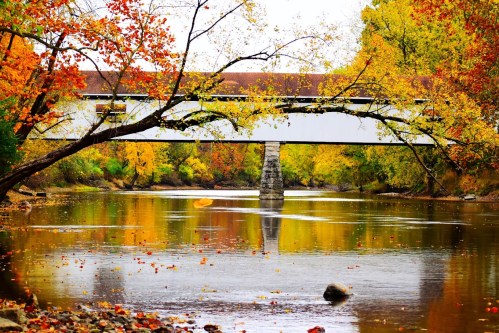 A picturesque scene of a covered bridge over a calm river, surrounded by vibrant autumn foliage.