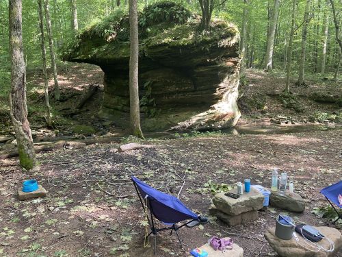 A serene forest scene featuring a large rock formation, a small stream, and camping gear set up nearby.