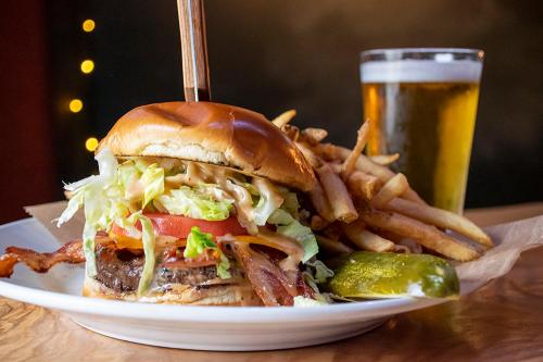 A juicy burger with bacon, lettuce, tomato, and pickles, served with fries and a glass of beer in the background.