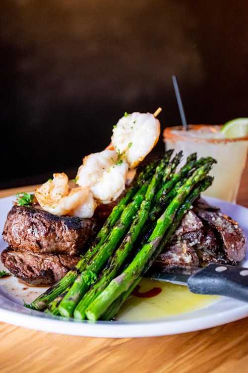 A plate featuring grilled steak, shrimp skewers, and asparagus, accompanied by a cocktail.