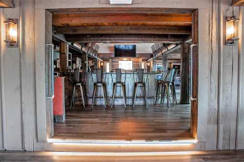 View of a rustic bar area with metal stools, wooden beams, and warm lighting, framed by a wooden entrance.