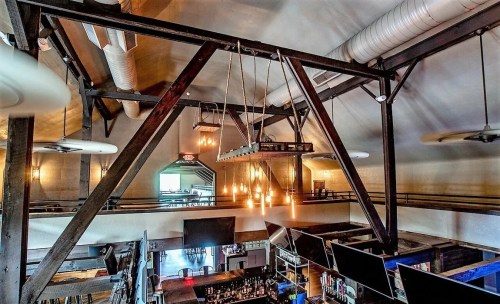 Interior of a stylish restaurant with high ceilings, wooden beams, and hanging lights, showcasing a bar area below.
