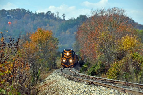 A train curves along a track surrounded by colorful autumn foliage and rolling hills under a blue sky.