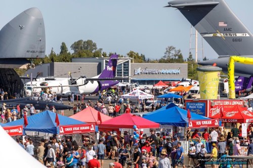 A busy airshow scene with crowds, food stalls, and various aircraft on display under a clear blue sky.
