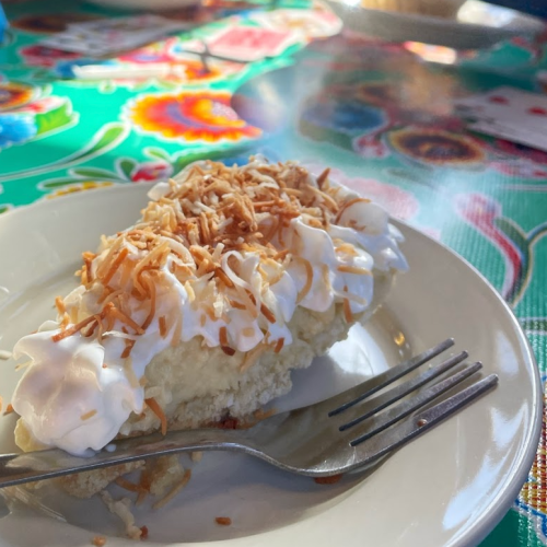 A slice of coconut cream pie topped with whipped cream and toasted coconut, served on a plate with a fork.