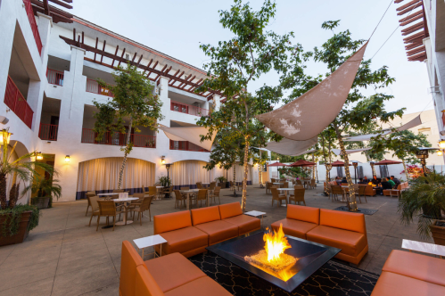 A cozy outdoor lounge area with orange seating, a fire pit, and greenery, surrounded by white buildings and shade sails.