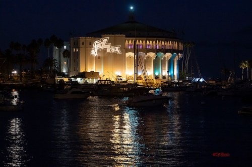A waterfront view of a brightly lit building at night, reflecting on the water with boats docked nearby.