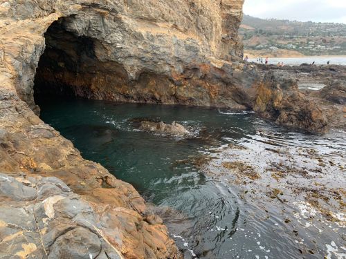 A rocky coastal cave with clear water inside, surrounded by rugged cliffs and a distant shoreline.