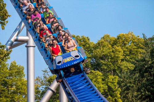 A roller coaster with excited riders descending a steep drop, surrounded by green trees under a clear blue sky.