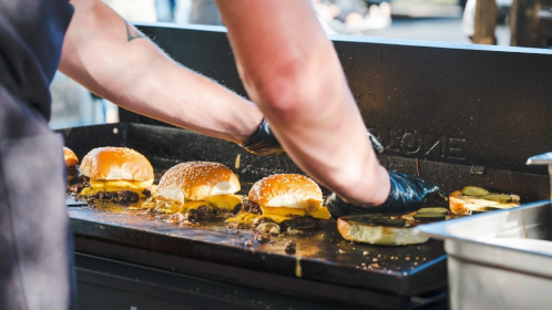 A person in gloves prepares burgers on a grill, with melted cheese and pickles on sesame seed buns.