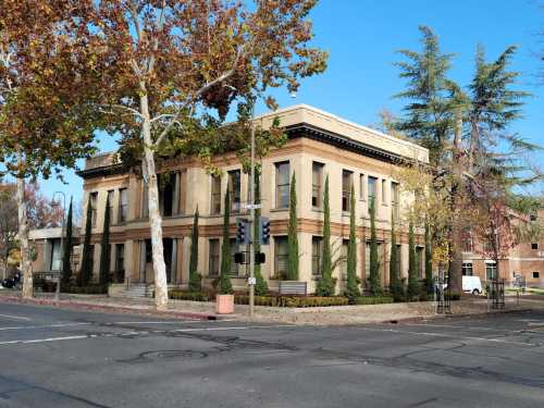 Historic building with tall trees and shrubs, located at a street corner under a clear blue sky.