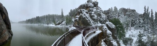 A snowy landscape featuring a winding boardwalk along a river, surrounded by rocky cliffs and evergreen trees.