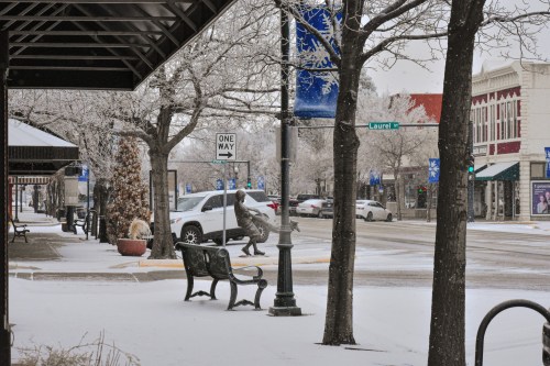 A snowy street scene with trees, a bench, and a statue, featuring a one-way sign and buildings in the background.