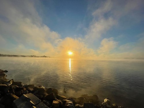 A serene sunrise over a misty lake, with soft clouds reflecting on the water and rocky shoreline in the foreground.