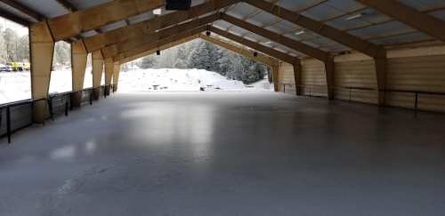 Indoor ice skating rink with a smooth, frozen surface and wooden beams overhead, surrounded by snow outside.