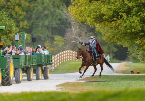 A knight in armor rides a horse alongside a wagon of people, waving a sword in a scenic outdoor setting.