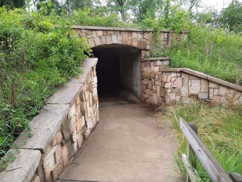 A stone archway leads into a dark tunnel, surrounded by greenery and a sloped pathway.