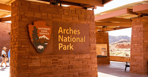 Sign for Arches National Park, featuring the National Park Service logo, with visitors walking in the background.