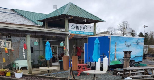 A rustic building with a teal roof, featuring a food truck and outdoor seating with blue umbrellas.