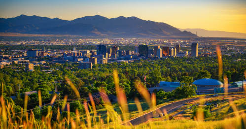 A panoramic view of a city skyline surrounded by mountains, with green fields in the foreground and a sunset glow.