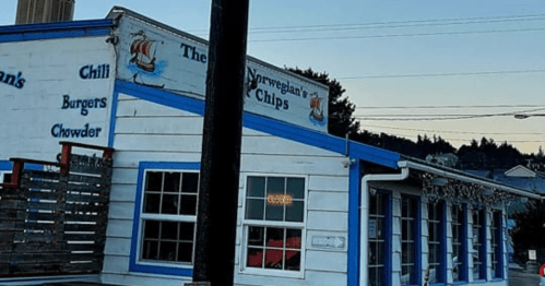A blue and white building with signs for "Chili, Burgers, Chowder" and "The Norwegian's Chips" at dusk.