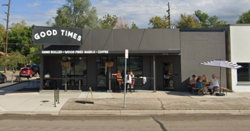 A modern café with a sign reading "Good Times," featuring outdoor seating and people enjoying bagels and coffee.