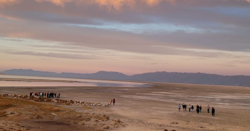 A group of people walking along a dry, sandy landscape with mountains and a colorful sky in the background.
