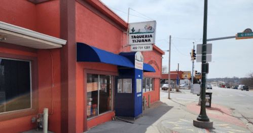 Exterior of a taqueria with a red building, blue awning, and a sign displaying the name and contact information.