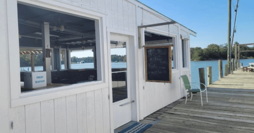 A waterfront seafood restaurant with a wooden deck, featuring large windows and a chalkboard menu outside.
