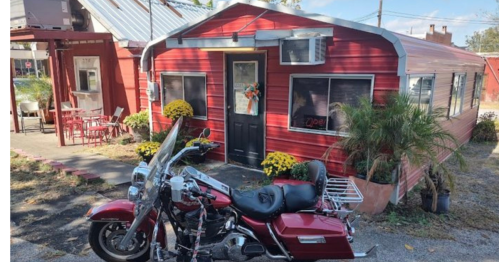 A red vintage-style building with a black door, surrounded by flowers, and a parked motorcycle in front.