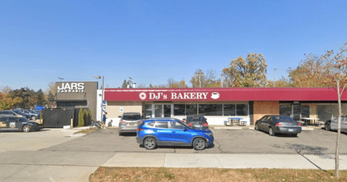 A bakery with a red awning and a sign reading "DJ's Bakery," next to a building labeled "JARS." Several cars are parked outside.