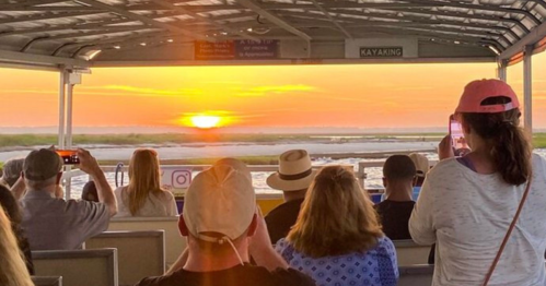 People on a boat watching a sunset, some taking photos, with a scenic view of water and land in the background.
