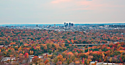 A panoramic view of a city skyline surrounded by vibrant autumn foliage in shades of orange, red, and yellow.