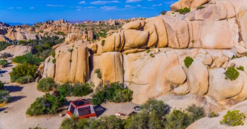 Aerial view of large rock formations and a small red tent in a desert landscape with shrubs and blue sky.