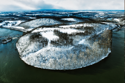 Aerial view of a snow-covered island surrounded by water, with trees and hills in the background under a cloudy sky.