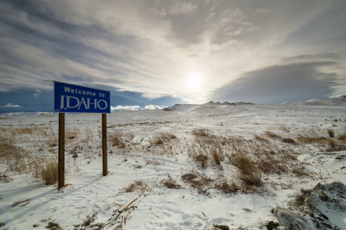 A "Welcome to Idaho" sign stands in a snowy landscape under a cloudy sky with the sun peeking through.