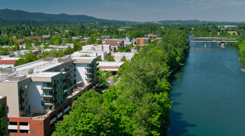 Aerial view of a river bordered by greenery and buildings, with hills in the background under a clear blue sky.