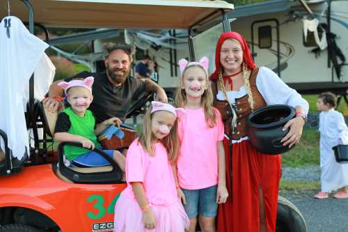 A group of children in costumes poses with an adult in a red riding hood outfit near a golf cart at a festive event.