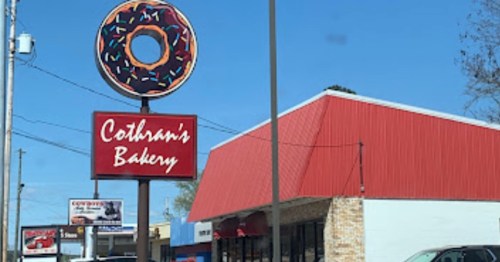 Sign for Cothran's Bakery featuring a donut with sprinkles, set against a clear blue sky and a red-roofed building.