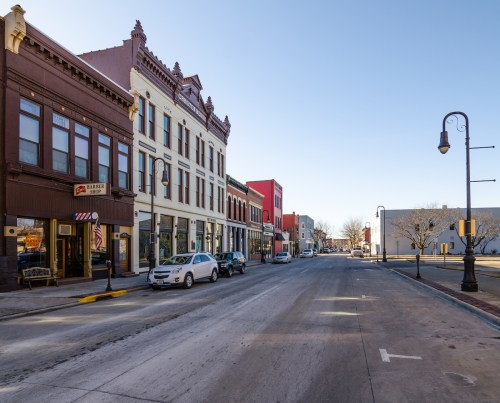 A quiet street lined with historic buildings, shops, and parked cars under a clear blue sky.