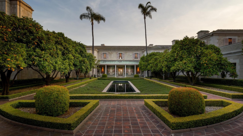 A serene courtyard with manicured hedges, palm trees, and a reflecting pool in front of a grand building at sunset.