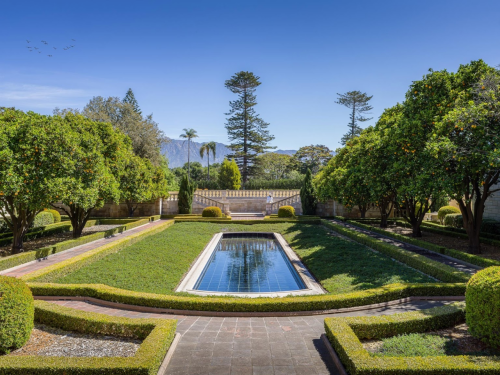A serene garden with neatly trimmed hedges, a reflecting pool, and trees under a clear blue sky.