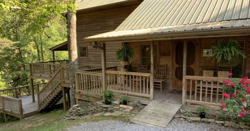 A rustic wooden house with a porch, surrounded by greenery and flowers, featuring a staircase and rocking chairs.